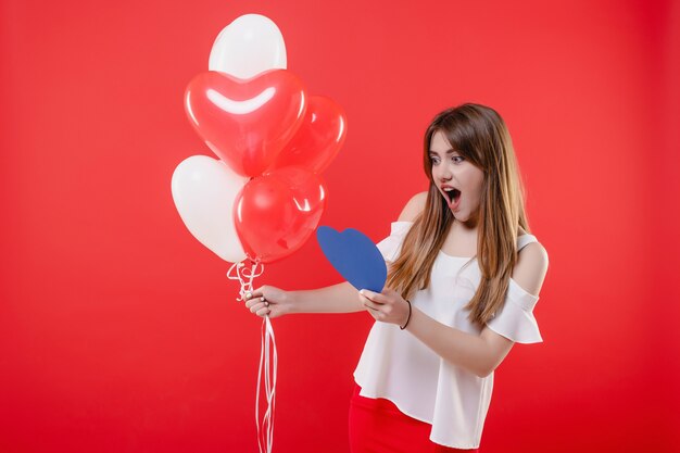 Femme avec carte de Saint Valentin en forme de coeur bleu et ballons colorés isolés sur mur rouge