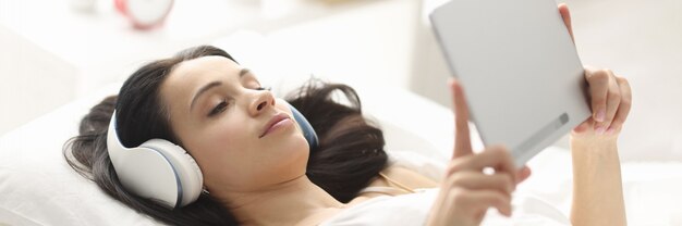 Femme avec un casque allongé sur le lit et tenant une tablette. Dommages causés par les gadgets avant le coucher