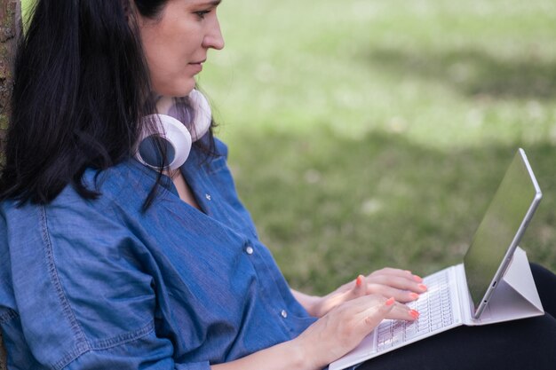 Femme casque sans fil à l'aide d'une tablette avec étui à clavier assis près d'un arbre dans le parc de printemps pigiste