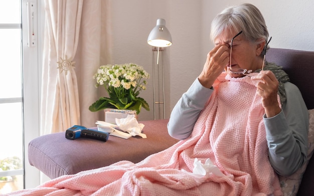 Femme caucasienne adulte âgée présentant des symptômes de maux de tête et de fièvre tels que la grippe saisonnière ou une allergie au pollen femme sur un canapé à la maison avec une couverture