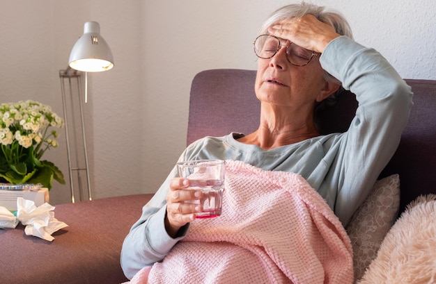 Femme caucasienne adulte âgée présentant des symptômes de rhume et de fièvre tels que la grippe saisonnière ou l'allergie au pollen tout en tenant un verre d'eau