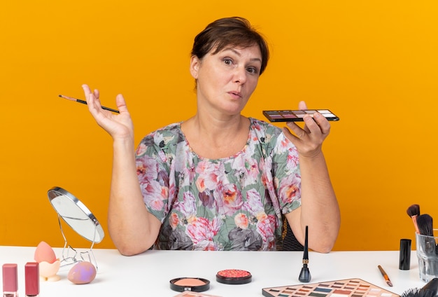 Femme caucasienne adulte surprise assise à table avec des outils de maquillage tenant une palette de fard à paupières et un pinceau de maquillage