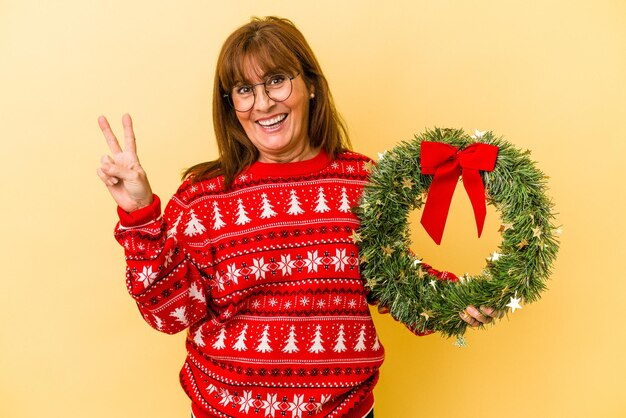 Femme caucasienne d'âge moyen célébrant Noël tenant une couronne isolée sur fond jaune joyeuse et insouciante montrant un symbole de paix avec les doigts.