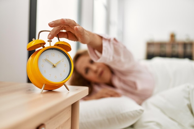 Femme caucasienne d'âge moyen éteignant le réveil allongé sur le lit dans la chambre.