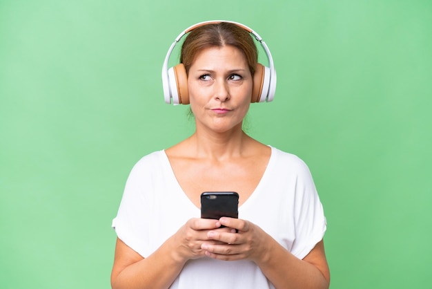 Femme caucasienne d'âge moyen sur fond isolé écoutant de la musique avec un mobile et pensant