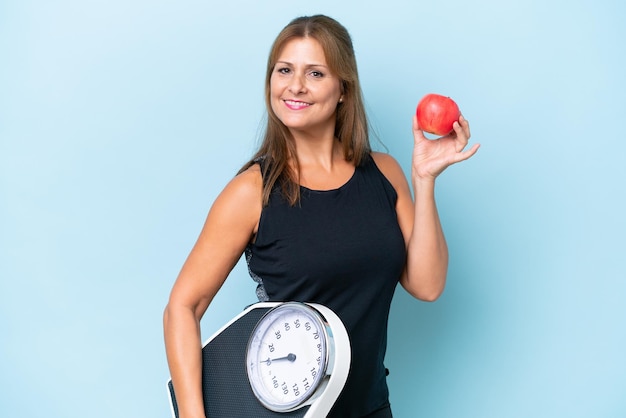 Femme caucasienne d'âge moyen isolée sur fond bleu avec balance et avec une pomme