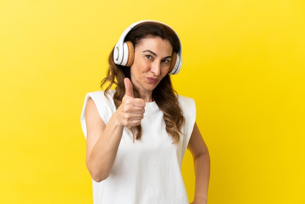 Femme caucasienne d'âge moyen isolée sur fond jaune, écoutant de la musique et avec le pouce vers le haut