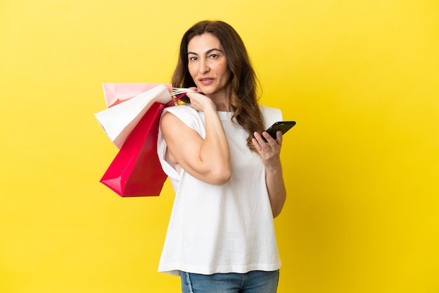 Femme caucasienne d'âge moyen isolée sur fond jaune tenant des sacs à provisions et un téléphone mobile