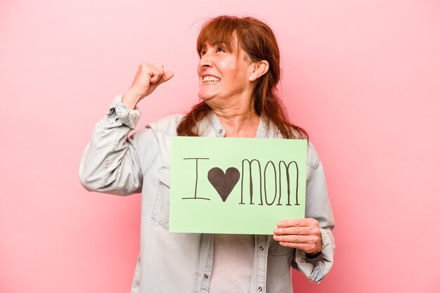 Femme caucasienne d'âge moyen tenant une pancarte j'aime maman isolée sur fond rose levant le poing après un concept de vainqueur de la victoire