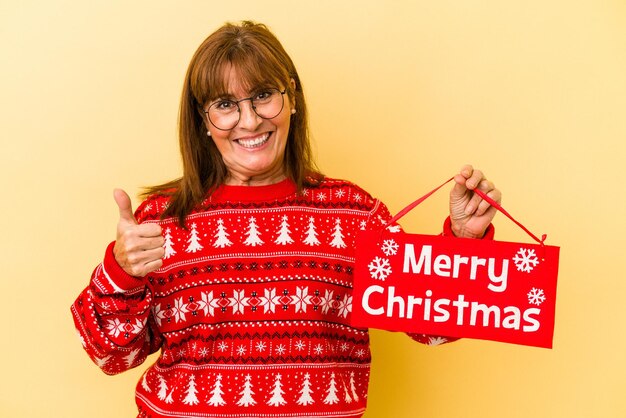 Femme caucasienne d'âge moyen tenant une pancarte "merry Christmas" isolée sur fond jaune souriant et levant le pouce vers le haut