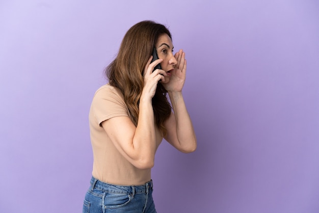 Femme caucasienne d'âge moyen utilisant un téléphone portable isolé sur fond violet criant avec la bouche grande ouverte sur le côté