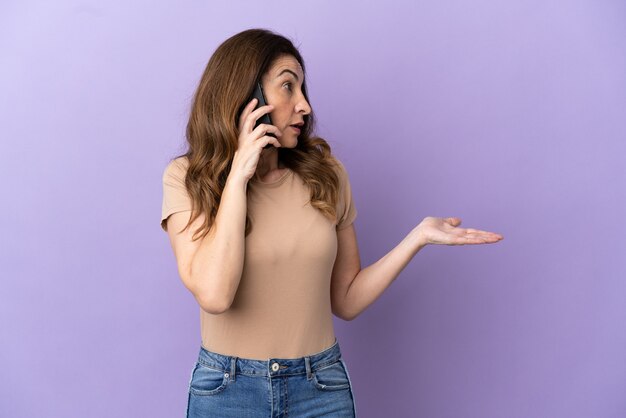 Femme caucasienne d'âge moyen utilisant un téléphone portable isolé sur fond violet avec une expression de surprise tout en regardant de côté