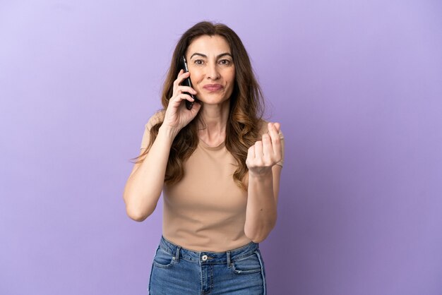 Femme caucasienne d'âge moyen utilisant un téléphone portable isolé sur fond violet faisant un geste d'argent