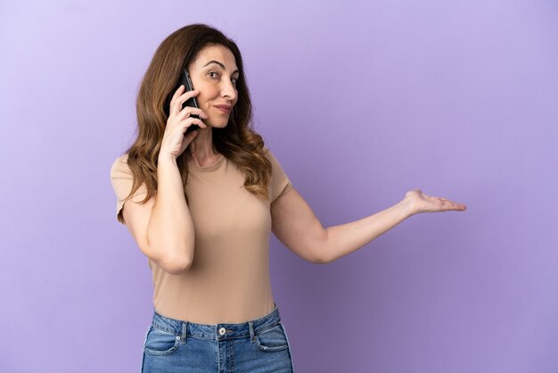 Femme caucasienne d'âge moyen utilisant un téléphone portable isolé sur fond violet tendant les mains sur le côté pour inviter à venir