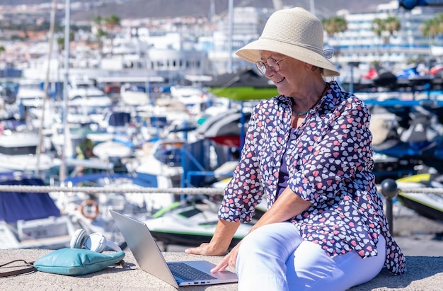 Femme caucasienne âgée avec chapeau assis à l'extérieur au port de mer à l'aide d'un ordinateur portable dans le travail à distance femme âgée aux cheveux gris naviguant avec un ordinateur