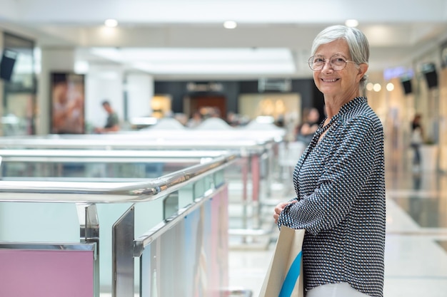 Femme caucasienne âgée debout à l'intérieur d'un centre commercial regardant en souriant à la caméra tenant un sac à provisions