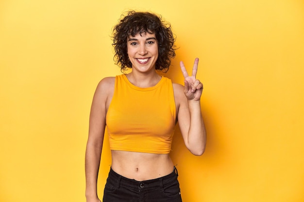 Une femme caucasienne aux cheveux bouclés en haut jaune joyeuse et insouciante montrant un symbole de paix avec des doigts