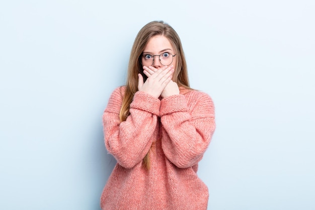 femme caucasienne aux cheveux rouges couvrant la bouche avec les mains avec une expression choquée et surprise, gardant un secret ou disant oups