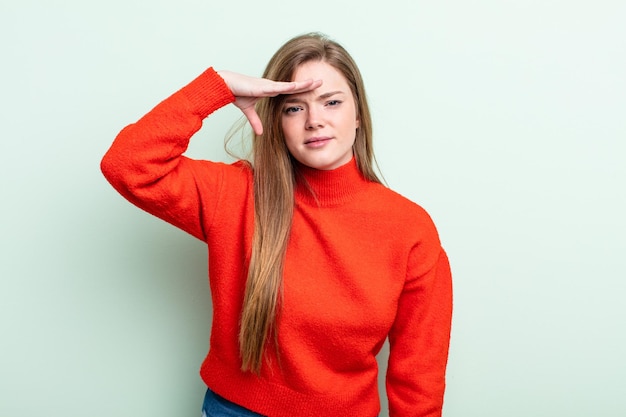 femme caucasienne aux cheveux rouges regardant perplexe et étonnée, la main sur le front regardant loin, regardant ou cherchant