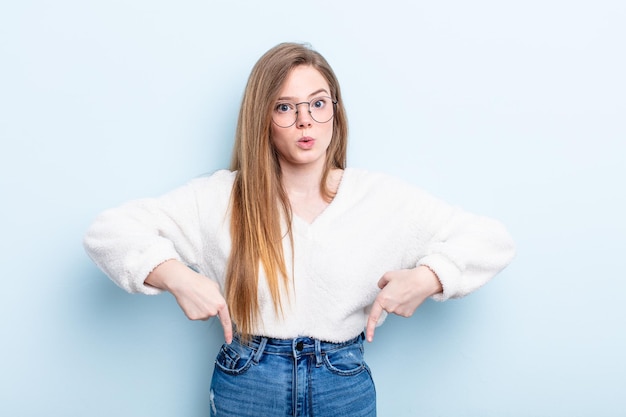 Femme caucasienne aux cheveux roux avec la bouche ouverte pointant vers le bas avec les deux mains semblant choquée, étonnée et surprise