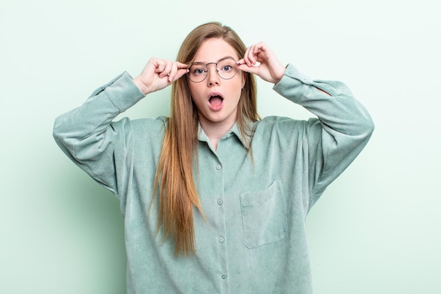 Femme caucasienne aux cheveux roux se sentant choquée, étonnée et surprise, tenant des lunettes avec un regard étonné et incrédule