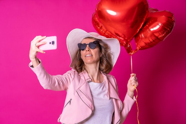 Une femme caucasienne blonde s'amusant avec un chapeau blanc et des lunettes de soleil dans une boîte de nuit avec des ballons cardiaques prenant un selfie avec son téléphone portable fond rose