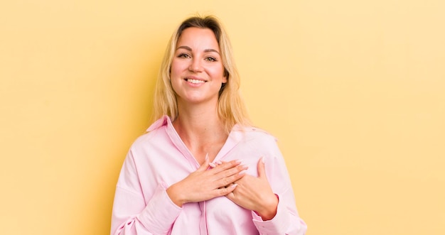 Femme caucasienne blonde se sentant romantique heureuse et amoureuse souriant joyeusement et se tenant la main près du cœur