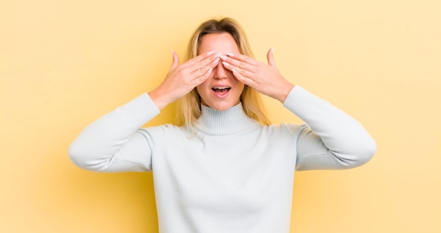 Femme caucasienne blonde souriante et se sentant heureuse couvrant les yeux avec les deux mains et attendant une surprise incroyable