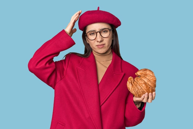 Une femme caucasienne chic mangeant un croissant en manteau rouge et un béret sur le fond du studio est choquée