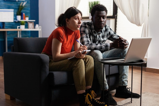 Femme caucasienne concentrée écrivant sur un ordinateur portable tandis qu'un homme afro-américain pensif l'aidait. Couple diversifié vérifiant les factures bancaires et la situation financière tout en discutant dans le salon à la maison.