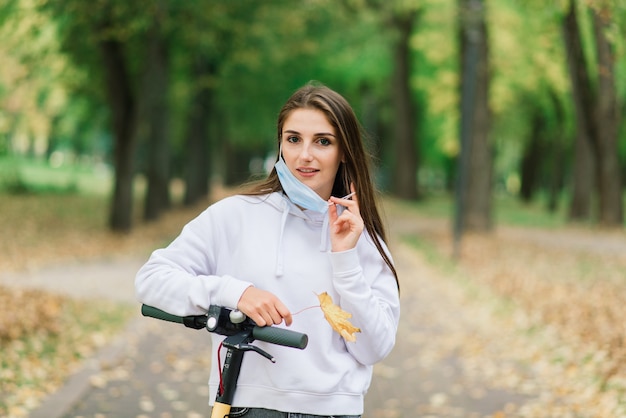 Femme caucasienne décontractée portant un masque protecteur faisant du scooter électrique urbain dans le parc de la ville pendant la pandémie de covid. Notion de mobilité urbaine.