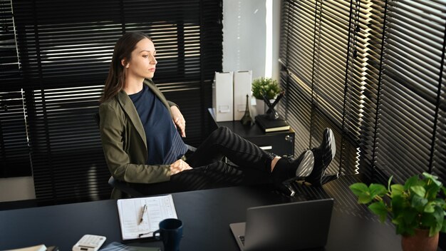 Femme caucasienne du millénaire satisfaite se relaxant sur le lieu de travail avec les jambes sur la table et regardant par la fenêtre du bureau