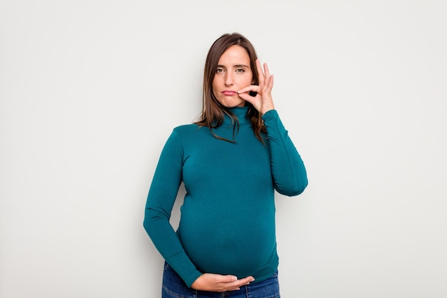 Une femme caucasienne enceinte isolée sur un fond blanc avec des doigts sur les lèvres gardant un secret