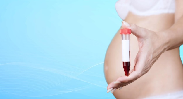 Une femme caucasienne enceinte en sous-vêtements tient un récipient en plastique pour recueillir le sang, un pot avec un bouchon rouge, un test sanguin, sur fond bleu. Fermer. Copiez l'espace. Photo de haute qualité