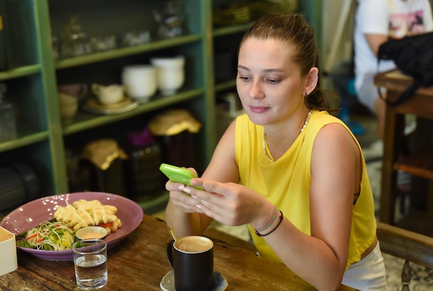 Une femme caucasienne fait défiler un téléphone et se prépare à prendre son petit déjeuner dans un café