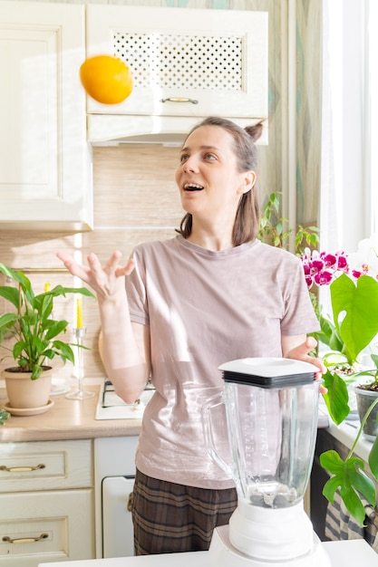 Une femme caucasienne heureuse et gaie en t-shirt dans sa cuisine fait un smoothie vert