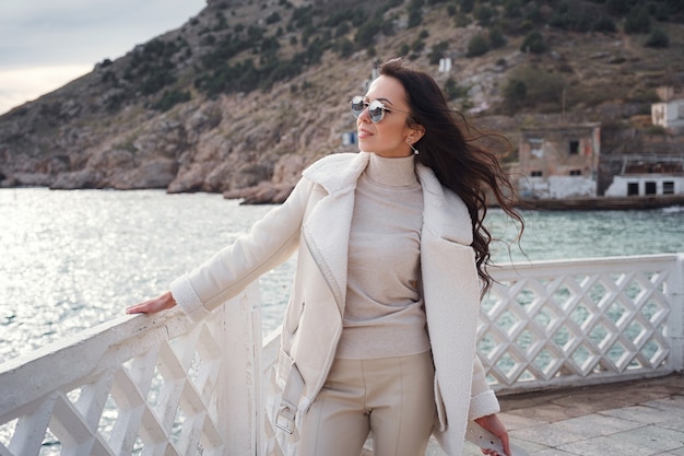 Une femme caucasienne insouciante vêtue de vêtements beiges profitant de la vue sur la mer par une journée chaude et venteuse. une femme portant des lunettes de soleil élégantes profitant d'une belle journée d'hiver et respirant l'air marin