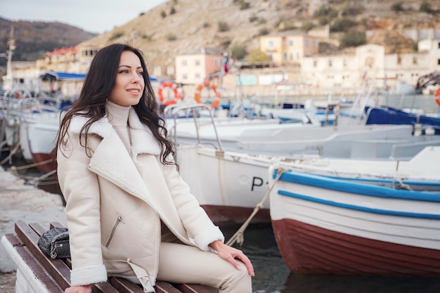 Photo une femme caucasienne insouciante vêtue de vêtements beiges profitant de la vue sur la mer par une journée chaude et venteuse. portrait devant une baie de mer avec des bateaux sur la jetée