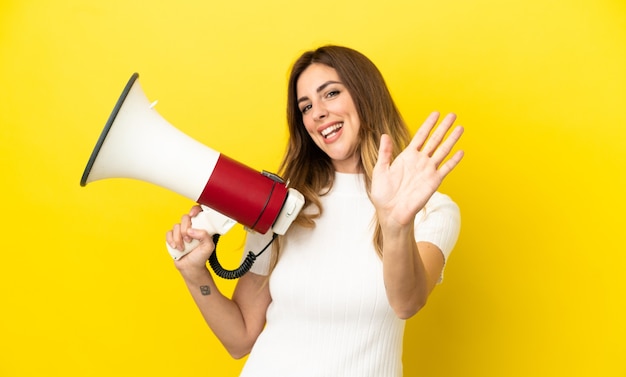 Femme caucasienne isolée sur une surface jaune tenant un mégaphone et saluant avec la main avec une expression heureuse