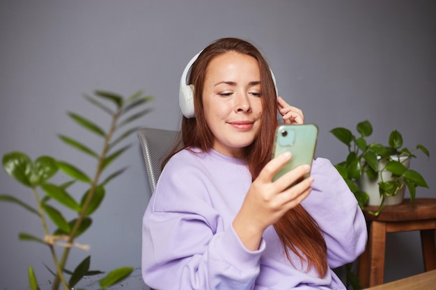 Femme caucasienne joyeuse écoutant de la musique ou un podcast et tenant un smartphone Beaucoup de plantes vertes autour d'une femme aux cheveux roux en pull violet