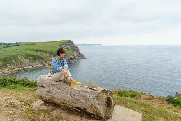 Une femme caucasienne joyeuse isolée s'assoit sur un banc de falaise. Vue horizontale d'une femme voyageant sur la côte. Concept de personnes et de voyage.