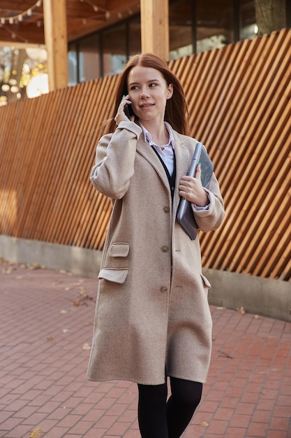 Femme caucasienne en manteau beige élégant appelant par smartphone tenant un ordinateur portable