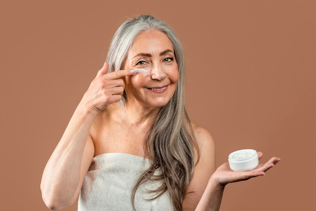 Une femme caucasienne mature souriante avec une beauté naturelle montre un pot et applique de la crème sur le visage recommande le rajeunissement