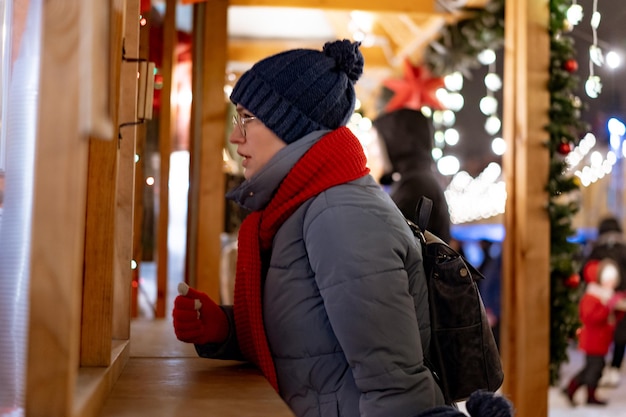 Femme caucasienne portant un chapeau et une écharpe rouge achetant des friandises en kiosque au marché de Noël