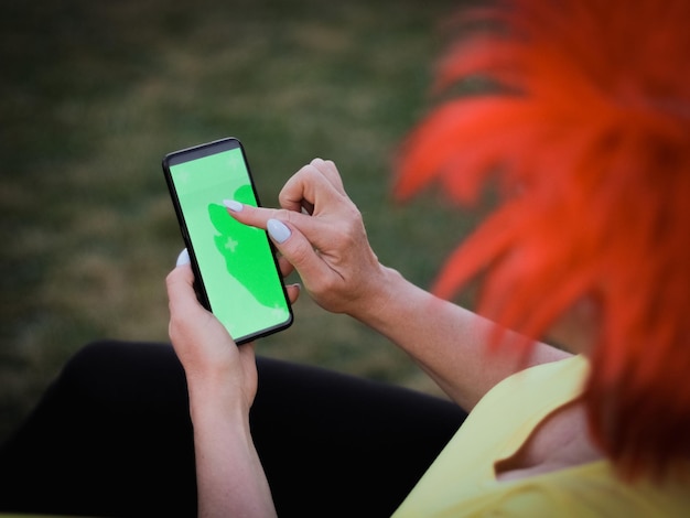 Femme caucasienne portant une perruque de drapeau belge tenant un smartphone avec un écran vert pointant son doigt dessus