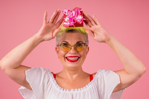 Une femme caucasienne porte des lunettes de soleil avec des cheveux teints en jaune élégants tenant une boîte cadeau en fond de studio rose.