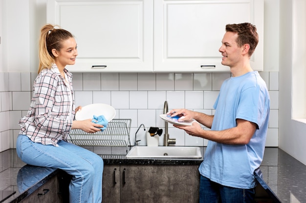 Femme Caucasienne Positive Essuyant La Vaisselle Pendant Que Son Mari Se Lave à La Cuisine