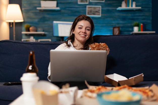 Femme caucasienne reposant sur un canapé regardant la caméra en train de manger un délicieux hamburger tout en travaillant sur un ordinateur portable