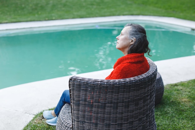 Femme caucasienne senior réfléchie assise sur une chaise au bord de la piscine dans le jardin. mode de vie à la retraite, passer du temps seul à la maison.