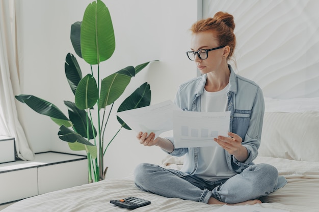 Une femme caucasienne sérieuse rousse est assise dans une pose de lotus sur des documents d'études de lit calcule les factures